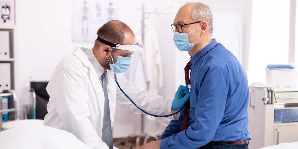A doctor wearing a face mask and shield uses his stethoscope to check
    the heart of a patient.
